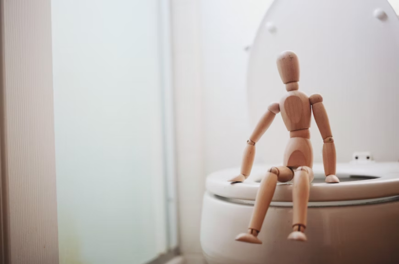 A brown wooden doll sitting on a white toilet bowl 