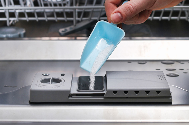 A hand tilts a scooper filled with dishwasher detergent into the unit slot
