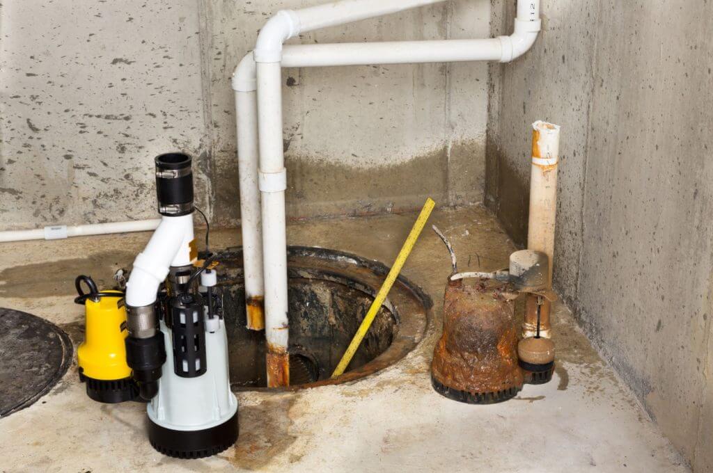 An opened sump pump pit with a yellow measuring stick plunged inside and canisters standing near its edges