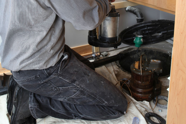 A crouched person holds a sump pump in front of an opened sump pit 